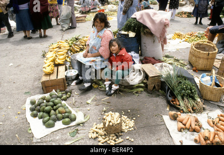 Lächelnde Frau und Kind sitzen auf kleinen Holzkiste inmitten von Obst und Gemüse. Lokalmarkt Hochland Ecuadors. Stockfoto