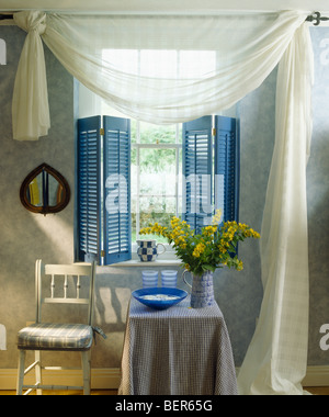 Weißen Voile Vorhänge und blauen Plantage Fensterläden am Fenster hinter kleinen Stuhl und Tisch im Ferienhaus Wohnzimmer Stockfoto