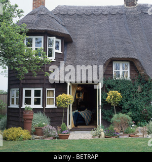 Clap-Tafel reetgedeckte Landhaus mit weißen Fenstern und abgeschnitten Bäume in Töpfen auf beiden Seiten der Front-Tür öffnen Stockfoto