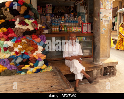 Ältere Ladenbesitzer sitzt vor seinem Supermarkt neben einem Haufen Garn zum Verkauf in Rajasthan, Indien Stockfoto