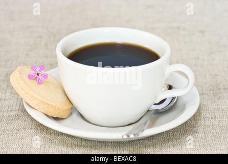 Eine weiße Tasse schwarzen Kaffee mit einem Shortbread-Keks und eine dekorative kleine Blume. Stockfoto