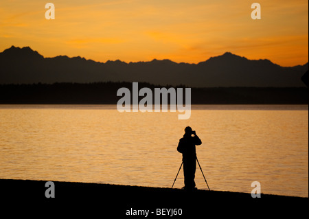 Ein Fotograf auf Alki Beach in West Seattle, Washington. Die Sonne geht über die Olympic Mountains und Bainbridge Island. Stockfoto