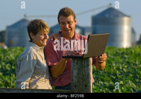 Mann und Frau Bauern in ihre Sojabohnen Feldeingaben schneiden Daten in ihrer Laptop-Computer mit Getreidesilos im Hintergrund. Stockfoto