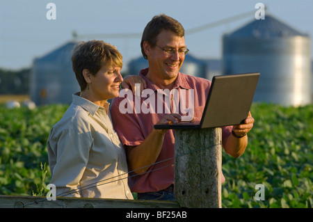 Mann und Frau Bauern in ihre Sojabohnen Feldeingaben schneiden Daten in ihrer Laptop-Computer mit Getreidesilos im Hintergrund. Stockfoto