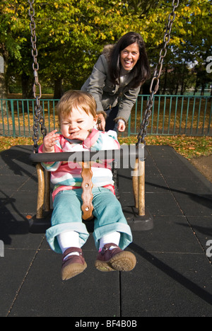 Vertikale Nahaufnahme Portrait von eine junge Mutter schob ihr kleines Mädchen auf einer Schaukel im Park. Stockfoto