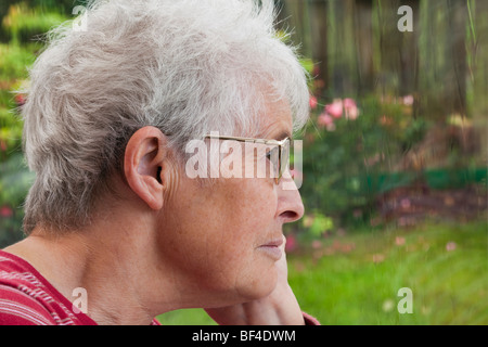 Ältere Seniorin OAP-Dame mit einem verzweifelten Ausdruck, die allein zu Hause an einem regnerischen Tag durch ein Fenster in den Garten blickt. Großbritannien Stockfoto