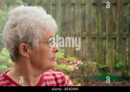 Ältere ältere Frau mit einem MUTLOS-Expression durch ein Fenster allein und fühlen sich durch einen regnerischen Tag deprimiert. UK psychische Gesundheit Depression Stockfoto