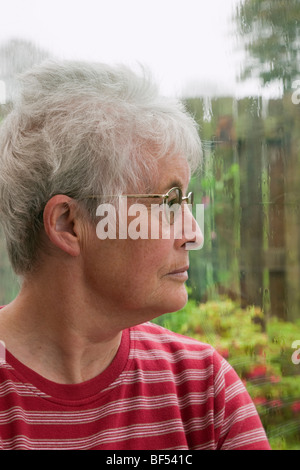 Ältere Frau mit einem MUTLOS-Expression durch ein Fenster an einem regnerischen Tag. Großbritannien Großbritannien Stockfoto