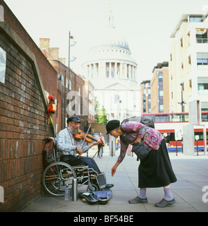 Eine Frau, die Geld geben, Nächstenliebe, ein Straßenmusikant spielt die Geige sitzt in einem Rollstuhl in der Nähe von St. Paul's Cathedral London KATHY DEWITT Stockfoto