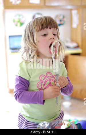 Junges Mädchen, drei Jahre alt, lecken den Quirl nach helfen, einen Kuchen zu mischen. Stockfoto