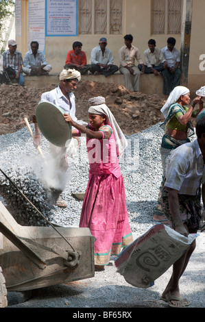 Junge indische Frau arbeiten auf den Straßen werfen Steine von einer Schüssel in ein Betonmischer. Puttaparthi, Andhra Pradesh, Indien Stockfoto