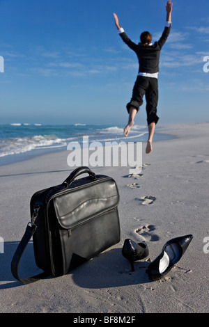 Frau im Büro Kleidung am Strand springen mit Freude und Freiheit. Laptop-Tasche und Schuhe im Vordergrund. Stockfoto