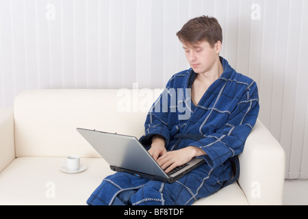 Der junge Mann arbeitet mit dem Laptop in einen blauen Morgenmantel Stockfoto