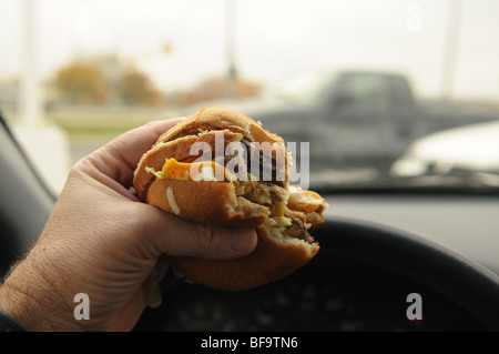 Ein Autofahrer hält einen Cheeseburger, wie er im Verkehr Essen fährt, eine Tätigkeit, die viele sagen ist eine gefährliche Ablenkung Stockfoto