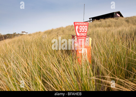 Rot Notfall Telefon in Dünengebieten Grass mit Rettungsschwimmer-Hütte im Hintergrund, Sennen, Cornwall, UK. Stockfoto