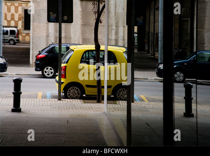 Ein gelbes Stadtauto in Girona, Spanien, spiegelt sich im Glas eines modernen Gebäudes wider und erzeugt eine amüsante optische Illusion eines seltsam geformten Fahrzeugs. Stockfoto