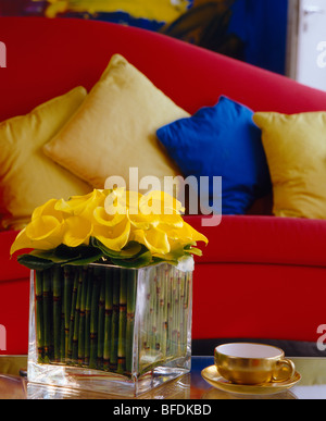 Gelbe Calla-Lilien in Glasvase auf Tisch vor roten Sofa mit gelben Kissen Stockfoto