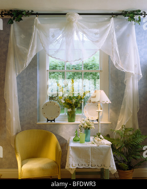 Weißen Voile Vorhänge auf Hütte Fenster über gelben Stuhl und einem kleinen Tisch mit Creme Lampe und weißes Tuch Stockfoto