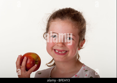 kleines Mädchen (7 Jahre alt), das vorderen Zähne zu essen Apfel auf rote Bohnenbeutelstuhl vor weißem Hintergrund fehlt Stockfoto