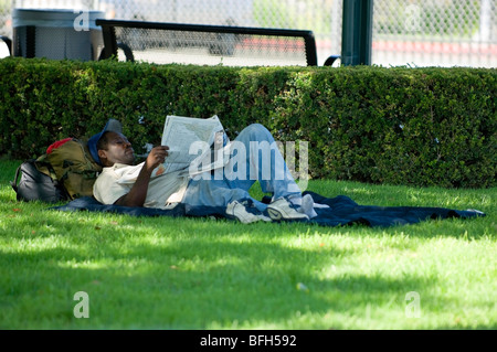 Mann liest Zeitung auf einer Rasenfläche beim stillstehen auf seinen Rucksack, Taschen oder Gepäck. Stockfoto