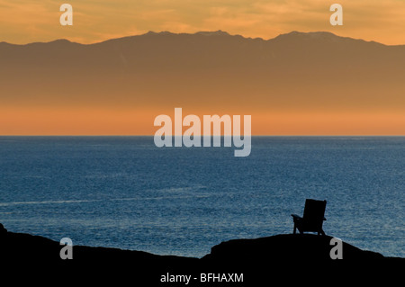 Ein Adirondack Stil Stuhl ist Silhouette gegen den Ozean und die Olympic Mountains bei Sonnenuntergang in der Nähe von Victoria BC. Stockfoto