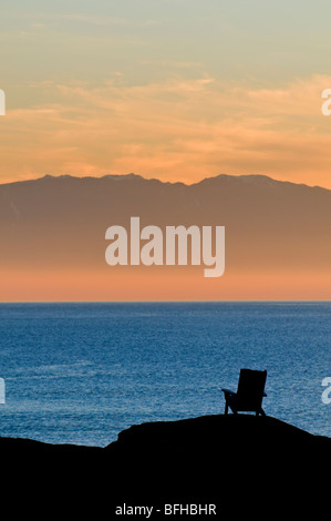 Ein Adirondack Stil Stuhl ist Silhouette gegen den Ozean und die Olympic Mountains bei Sonnenuntergang in der Nähe von Victoria BC. Stockfoto
