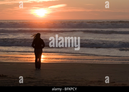 Eine anonyme Frau gesehen im Silhouette stehen am Strand bei Sonnenuntergang, ihr Haar im Wind wehen. Stockfoto