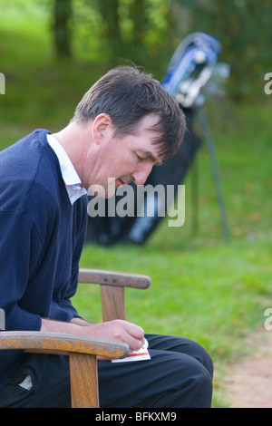 Ein im mittleren Alter weißer Mann Golfen nach Abschluss ein Loch mit seinem Golf-Trolley im Hintergrund, seine Scorekarte ausfüllen Stockfoto