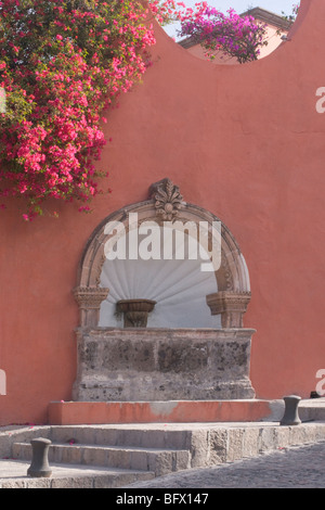 Bogen Sie geformte Brunnen in einem Adobe Terrakotta Wand, rosa Bougainvillea. Graue Treppe führt diagonal von Straße mit Kopfsteinpflaster. Stockfoto