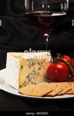 Keil von Stilton, begleitet von einem Glas roten Wein Cracker und Reife strauchtomaten auf einem weißen Teller vor schwarzem Hintergrund. Stockfoto