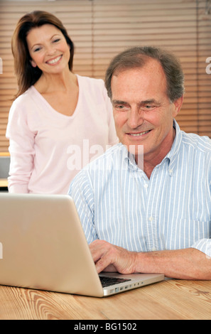 Lächelnde Frau schaut Ehemann oder Partner, wie er einen silbernen Laptop-Computer am Küchentisch benutzt Stockfoto