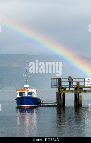 Kleine blaues Boot vertäut am Luss Pier am Loch Lomond mit Regenbogen im Hintergrund Stockfoto