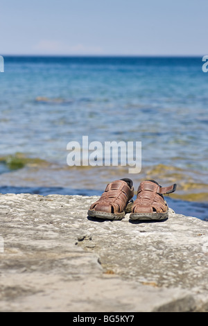 Ein paar braune Sandalen auf einem Felsvorsprung, mit einer Fläche von blauem Wasser und Himmel dahinter. Stockfoto