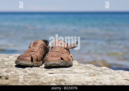 Ein paar braune Sandalen auf einem Felsvorsprung, mit einer Fläche von blauem Wasser und Himmel dahinter. Stockfoto