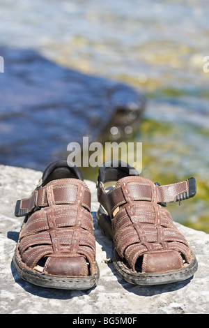 Eine Nahaufnahme von ein paar braune Sandalen auf einem Felsvorsprung, mit einer Fläche von Wasser zurück. Stockfoto