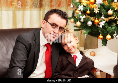 Vater umarmt Sohn neben dem Weihnachtsbaum lächelt und schaut in die Kamera Stockfoto