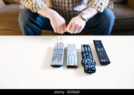 Mann sitzt auf der Couch, mit Fernbedienungen, aufgereiht auf einem Couchtisch. Stockfoto