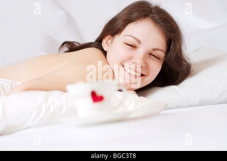 glückliche Frau im Bett und Tasse mit Herz vor ihr Stockfoto