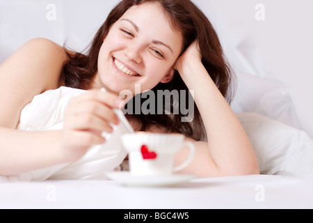 glückliche Frau im Bett und Tasse mit Herz vor ihr Stockfoto