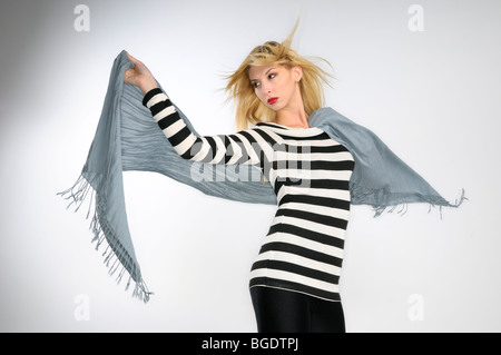 Junge, blonde Frau in gestreiften Oberteil hält einen Schal in den Wind auf grauem Hintergrund Stockfoto