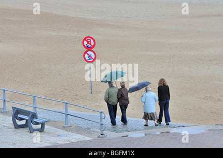 Touristen mit Sonnenschirmen zu Fuß über den Deich entlang Strand an einem regnerischen Tag in den Sommerferien an der belgischen Küste Stockfoto