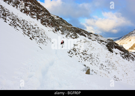 Eine Hügel Walker kreuzt ein Schneefeld am Unterlauf der Snowdon Pyg Track bei winterlichen Bedingungen. Stockfoto