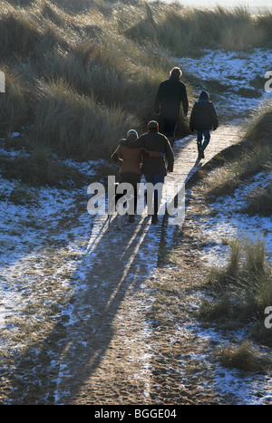 Vier Menschen und zwei Hunde, die zu Fuß auf einem Holzsteg durch verschneite, frostigen Dünen am Silvester Tag 2010 am Burnham Overy. Stockfoto