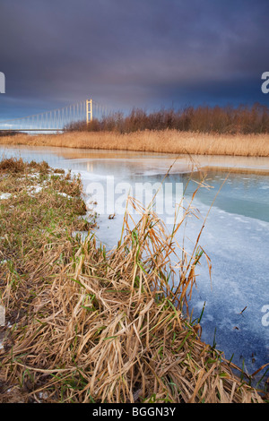 Ein Gefrorener Teich mit Schilf im Vordergrund und die Humber-Brücke in der Ferne an einem kalten Wintertag Stockfoto