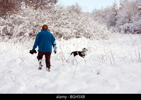 Dame in blauem Vlies spielt im Schnee, während ihr tief verschneiten Labradoodle Welpen beobachtet und wartet, dass ihr darauf Stockfoto