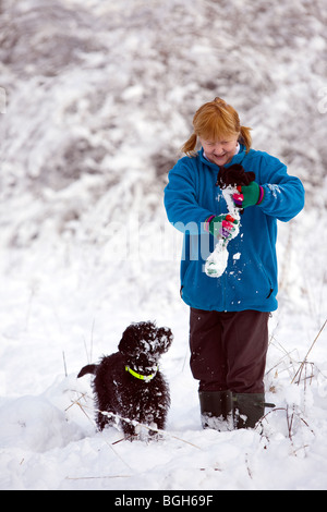 Dame in blauem Vlies wischt den Schnee von einem Hundespielzeug, während ihr tief verschneiten Labradoodle Welpen beobachtet und wartet Stockfoto