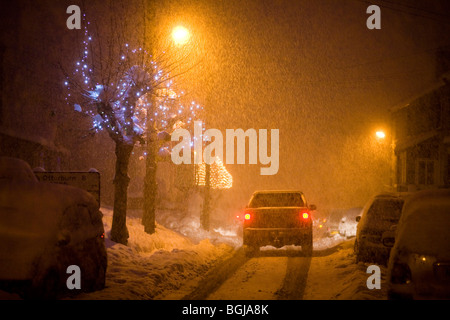 Auto fährt hinunter eine schneebedeckte Straße in der Nacht. Straßenbeleuchtung und Weihnachtsbeleuchtung Leuchten auf der Straße Stockfoto