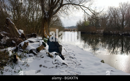 Mann mittleren Alters sitzen an Gillingham Strand am zugefrorenen Fluss Waveney mit Schnee auf dem Boden im winter Stockfoto