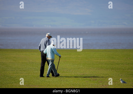 Alter Mann zu Fuß mit verbundenen Armen, mit einer alten Frau mit einem Stock am Meer bei Silloth, Cumbria. Stockfoto