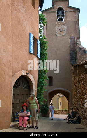 Glockenturm und Maler zeichnen Ocker gefärbt Häuserfassaden mit Sonnenrollos in Gasse, Roussillon, Provence, Vaucluse, Frankreich Stockfoto